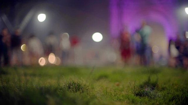 Green grass. A colourful crowd mowing at the background. People walking around. Creative evening lights and blurred shadows moving in the twilight. Festival in Barcelona.