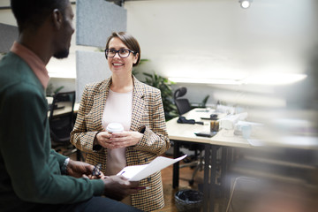 Waist up portrait of young business manager smiling cheerfully at African-American colleague while chatting at break in office, copy space
