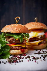 Two big classic american cheeseburger with two chops. Cheeseburgers on a linen tablecloth, decorated with different types of pepper and lettuce. Dark wooden background.