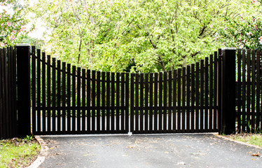 Dark wooden driveway property entrance gates set in timber picket fence with garden shrubs and trees in background