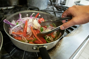 Line cook preparing peppers and onions with tongs and sauté pan on restaurant range
