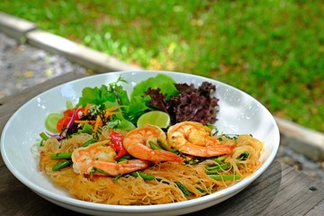 Fried Spicy Rice Vermicelli With Shrimp and Water Minosa in white plate on wooden table with green grass background.