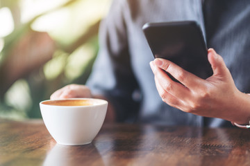 Closeup image of a woman holding  and using mobile phone while drinking coffee on wooden table in cafe