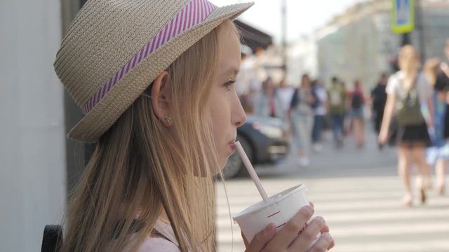 Young girl is drinking cocktail through a straw, standing on the street in city.