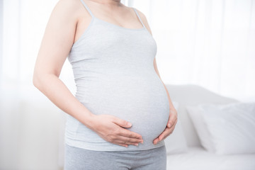 Pregnant woman standing and holding her belly by hands in bed room. Pregnancy, maternity, preparation and expectation concept.