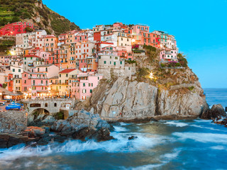 Colorful pastel houses along the cliffside in Manarola, Cinque Terre taken during the sunset with waves crashing along the coastline and a blue sky.