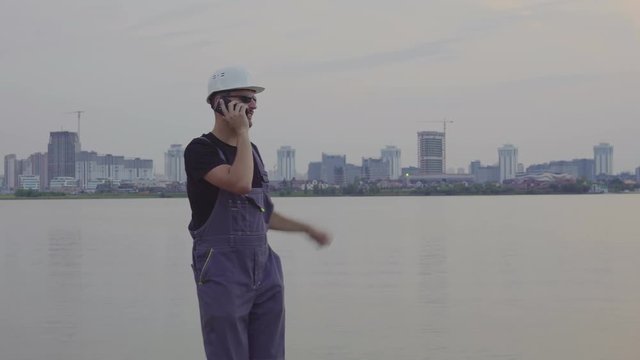 4K shot of handsome caucasian worker in white helmet talking on mobile phone near the beautiful evening city and construction site. construction success