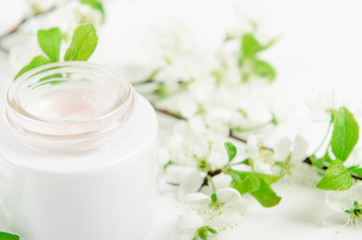 Fototapeta na wymiar Face cream in white jar on a white background with white small flowers of an apple tree. Concept natural cosmetics, organic beauty, delicate floral composition.