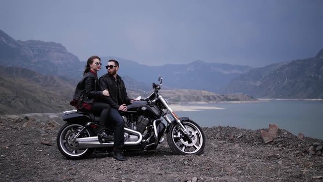 wonderful couple girl and guy sit on modern black and silver motorbike against calm lake among hills in evening