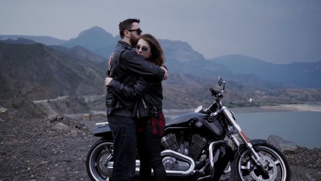 stylish couple in love hugs against black silver motorcycle and calm lake surrounded by mountains slow motion