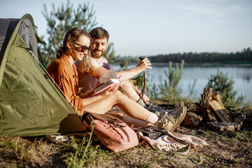 Young and cheerful couple having a picnic at the campsite while traveling in the forest near the lake during the sunset