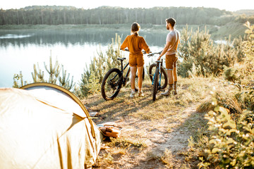 Young couple standing with mountain bicycles at the campsite, traveling in the forest near the lake...