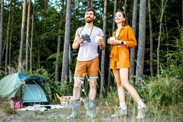 Young couple standing with hot drinks at the camping in the forest, enjoying their summer trip during the sunset