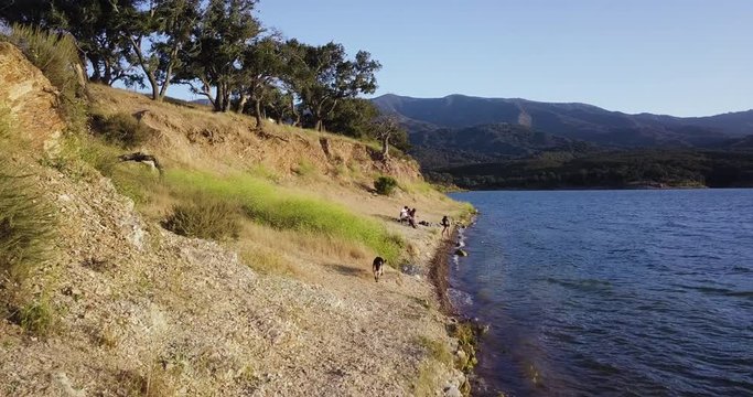 drone dolly in shot of a dog, german shepherd who is approaching his family sitting and relaxing on the shore of lake cachuma,California,USA