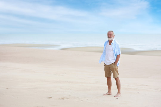 full shot image of older caucasian men, white beard and white hair in white and blue shirt is resting and standing on sandy beach after retirement in resort during the summer with left copy space