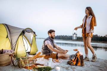 Young couple having fun at the campsite on the beach near the fireplace in the evening - obrazy, fototapety, plakaty
