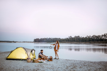 Young couple having fun at the campsite on the beach, standing near the fireplace during the evening. Wide landscape view