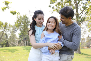 Medium shot of happy asian family, father, mother and daughter laughing . Happily parents hugging their little girl with smiling during summer time in the park