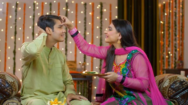 Young good looking brother and sister celebrating Raksha Bandhan / Bhai Dooj festival. Smiling Indian sister putting tika on her brother's forehead with pooja thali while celebrating Rakhi / Bhai Dooj