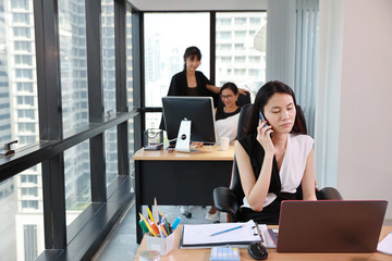 front view asian secretary woman or businesswoman wearing black and white dress working on laptop computer and cell phone in modern office with colleagues and city view background