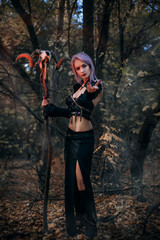 Naklejka premium A witch with a staff from the skull of a ram conjures in the forest. A girl in a black dress and purple hair. Image for halloween