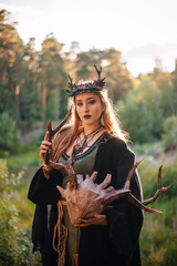 A girl in a terrible mask with horns is standing in nature. A man in a black cloak. The pagan witch conjures. Fantasy image