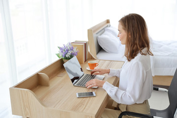 high angle view of beautiful caucasian girl drinking a cup of coffee or tea in the morning while working on laptop computer and cell phone on wood table in bedroom