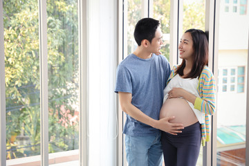 young asian husband standing and embracing, eyes contract together with young pregnant belly wife with happy smiling face and green trees background