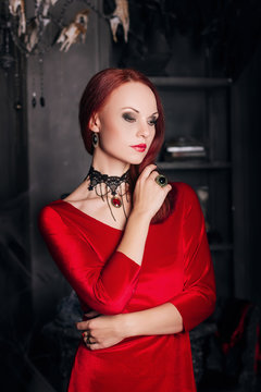 Beautiful red-haired girl with red lips. A model in a red dress with a black necklace in the castle. Vampire style. The image for halloween. Fantasy.