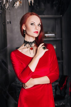 Beautiful red-haired girl with red lips. A model in a red dress with a black necklace in the castle. Vampire style. The image for halloween. Fantasy.