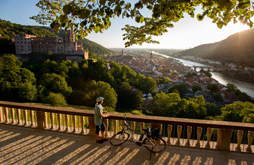 Fahrrad Fahrerin mit E-Bike blickt von der Scheffelterrasse in Heidelberg auf Schloss, Altstadt und alte Brücke, im Sonnen Abendlicht - obrazy, fototapety, plakaty
