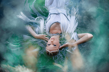 A girl elf in a white dress is floating in the water. Model in a medieval dress with pigtails...