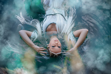 A girl elf in a white dress is floating in the water. Model in a medieval dress with pigtails floating in the lake