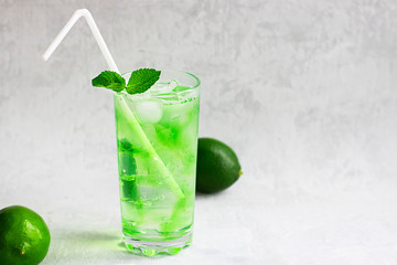 Summer cold green drink with lime, mint and lemon in tall glass with ice and straw and defocused fruit around on grey background. Sparkling beverage, alcohol drink, mojito coctail. Horizontal