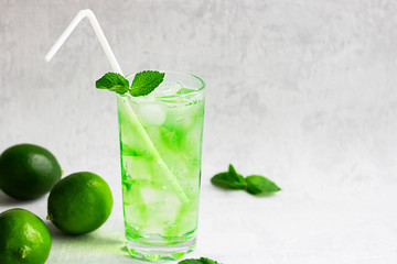 Summer cold green drink with lime, mint and lemon in tall glass with ice and straw and defocused fruit around on grey background. Sparkling beverage, alcohol drink, mojito coctail. Horizontal