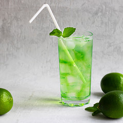 Summer cold green drink with lime, mint and lemon in tall glass with ice and straw and defocused fruit around on grey background. Sparkling beverage, alcohol drink, mojito coctail. Square