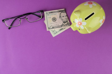 Green piggy bank and money and glasses on a bright purple background. Finance, savings, money. place for text. top view
