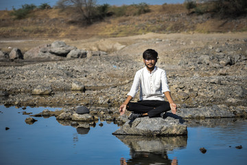 Portrait of fit and young Indian man doing meditation while sitting on rock at calm lake