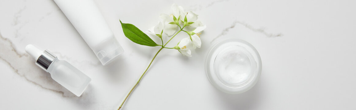 panoramic shot of jasmine, cosmetic glass bottle, jar with cream and moisturizer tube on white surface