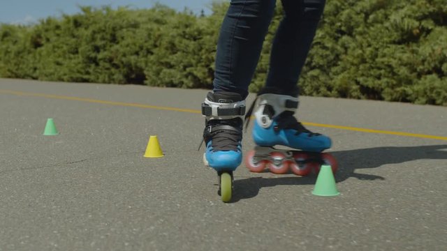 Close-up of female legs in roller blades riding forward crisscross through cones during workout in public park. Woman inline skating stunt of crisscross ride round cones standing in line on alley.