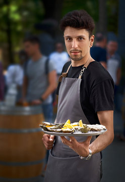 The waiter in an apron, offers a tray with fresh oysters on the ice with lemon, street trading on the street. Close up, appetizer to wine, healthy food