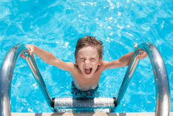 happy delightful small kid with wet hair standing on chrome handrail in fresh water swimming turquoise outdoors pool in sunny summer holiday leisure day with copy space