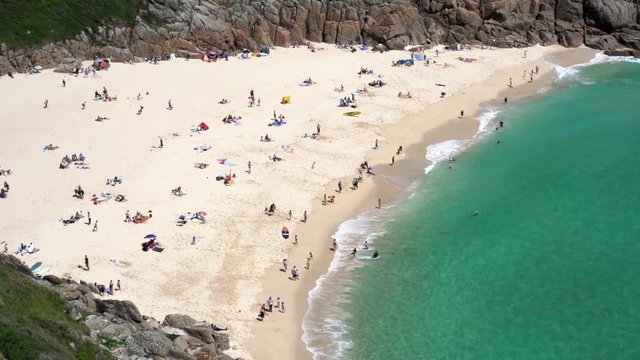Aerial view of Porthcurno Beach in Cornwall full of tourists, swimmers, surfers and families having fun on a beautiful spring sunny day. Zoom in 4k footage