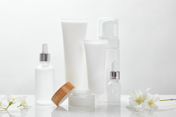 Fototapeta na wymiar jasmine flowers on white surface near glass bottles, cream in tubes, jar and cosmetic dispenser