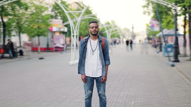 Zoom-out time lapse of attractive young man Arab standing in busy street with backpack looking at camera with serious face. Life, people and society concept.