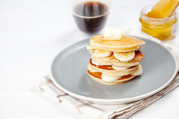 Closeup on fresh, homemade pancake stack with banana, melting butter and pouring honey or marple syrup. american pancake with glass of coffee on white background with morning light.