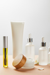 mascara bottle with natural oil, cream tube, cosmetic glass bottles, open jar with wooden cap and jasmine flower on beige