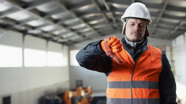 man beard and mustache in a hard hat and overalls is standing in the middle of the hangar and thumbs down at the camera