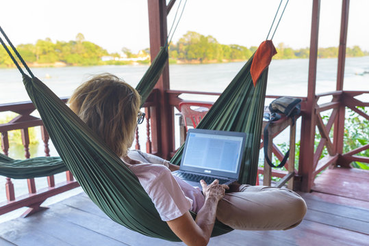 Woman working with laptop on hammock hanging in tourist resort on the Mekong River, Laos. Concept of millenials working around the world and digital nomads.
