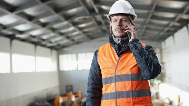 man beard and mustache in a hard hat and overalls is standing in the middle of the hangar and talking by his phone at the camera
